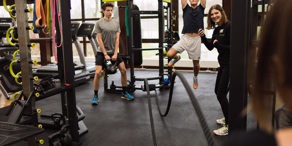 Trainerin mit Stoppuhr feuert 3 Trainierende am Functional Tower des Fitnessstudios an. 