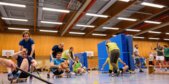 Teilnehmende des Kurses "Spiele spielen" spielen ein Sportspiel, sitzend auf Rollbrettern und mit Hockeyschlägern in den Händen.