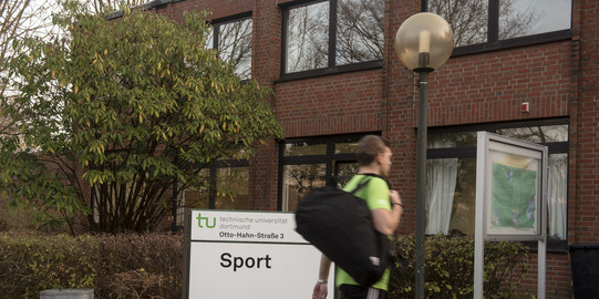 Person geht am Schild "Sport" vorbei in das Sportgebäude