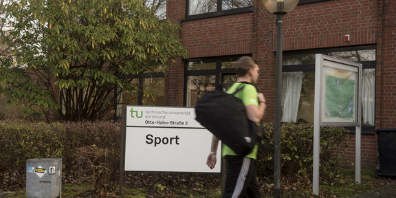 Person geht am Schild "Sport" vorbei in das Sportgebäude